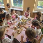 Dziewczynki w różowych fartuszkach siedzą przy stoliku i kroją owoce.