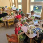 Chłopcy w zielonych fartuszkach siedzą przy stolikach, kroją owoce i wrzucają do słoików.