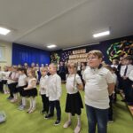Dzieci stoją z przodu ubrani na galowo, mówią wiersze do mikrofonu.