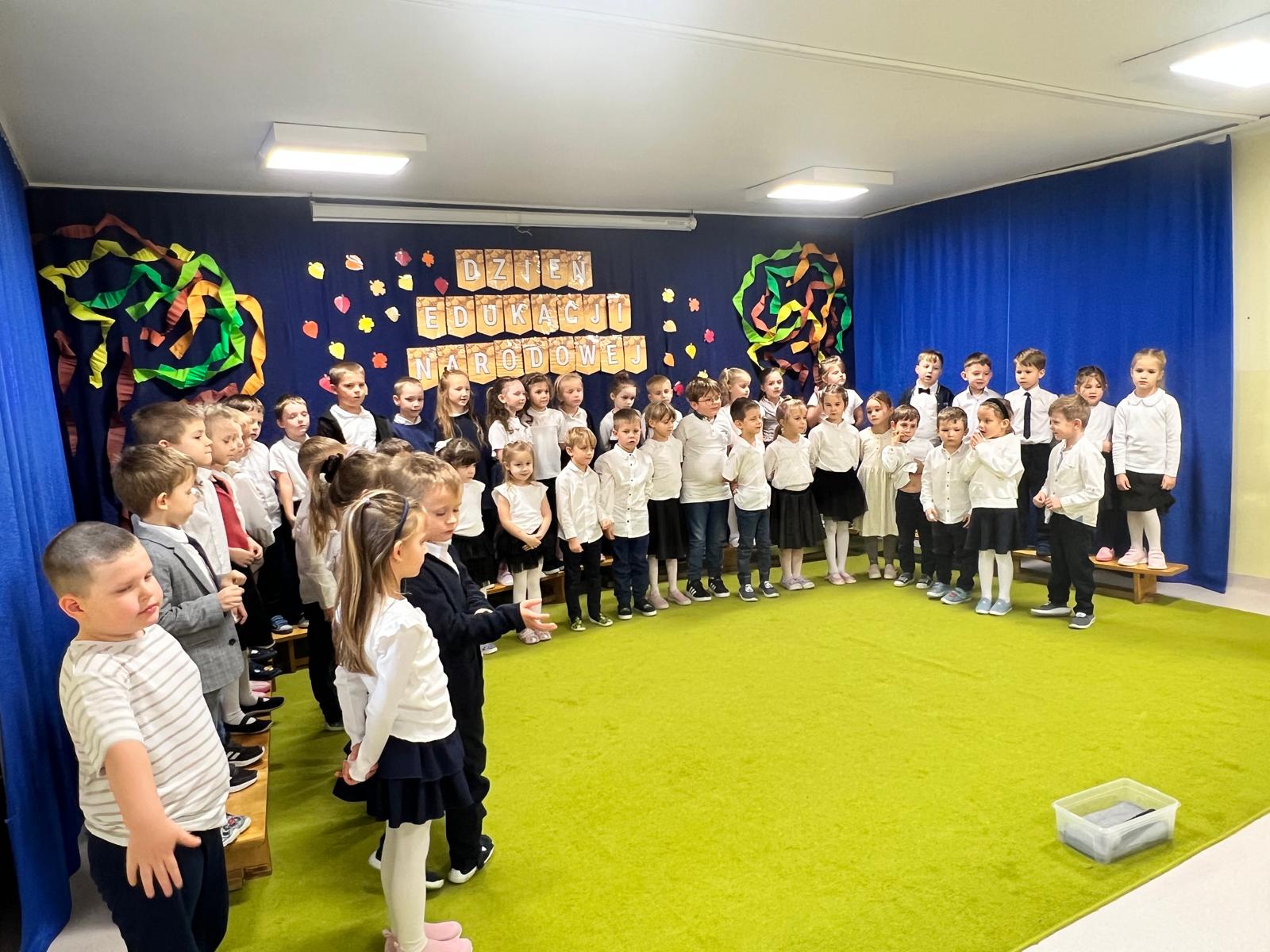 Przedszkolaki w galowych strojach stoją w sali udekorowanej z okazji Dnia Edukacji Narodowej.
