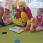 Kubuś Puchatek siedzi na dywanie z dziećmi i czyta im książkę.
