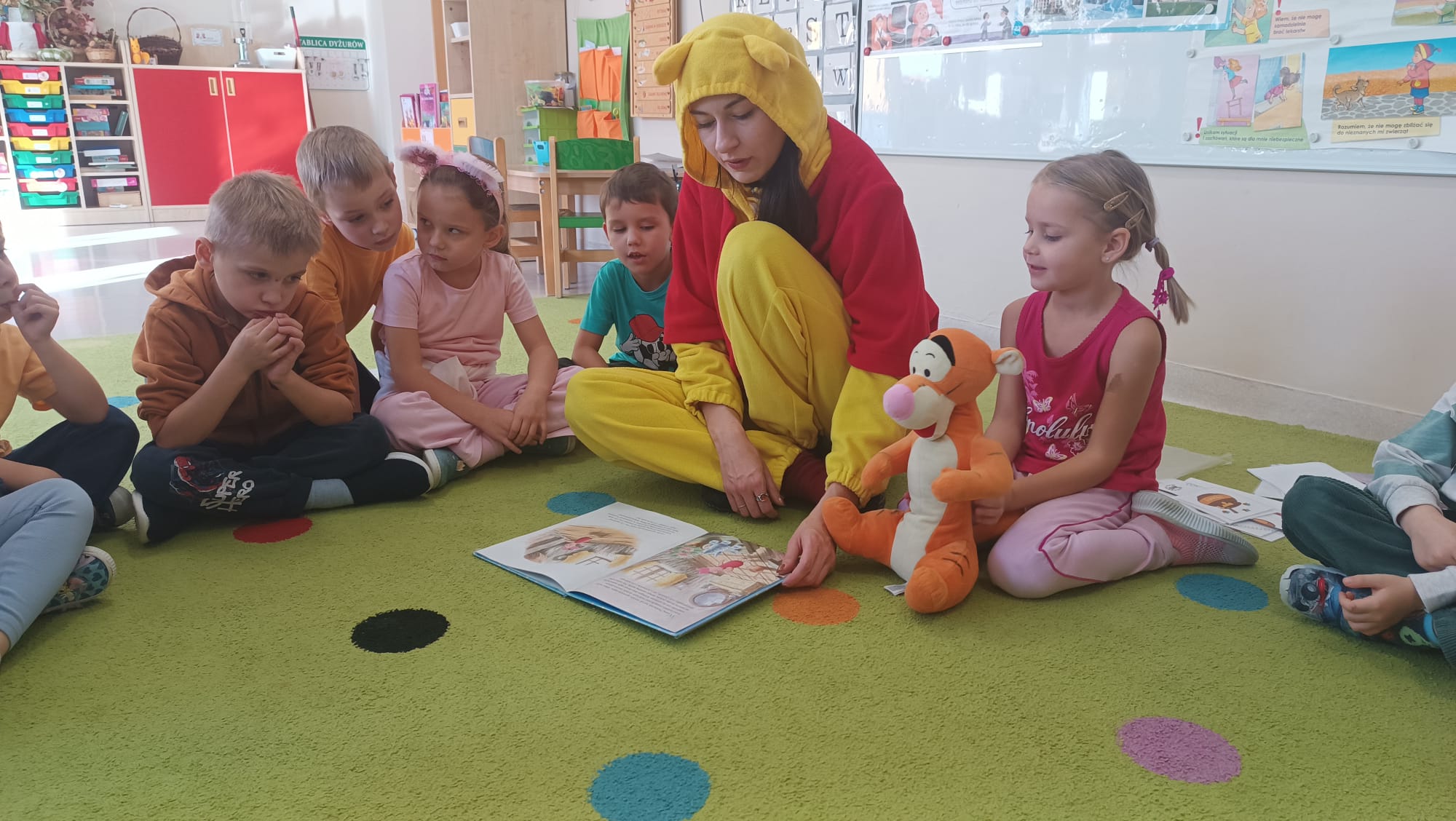 Kubuś Puchatek siedzi na dywanie z dziećmi i czyta im książkę.