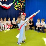 Dziewczynka ubrana w białą sukienkę, biało czerwony wianek macha Polską flagą.