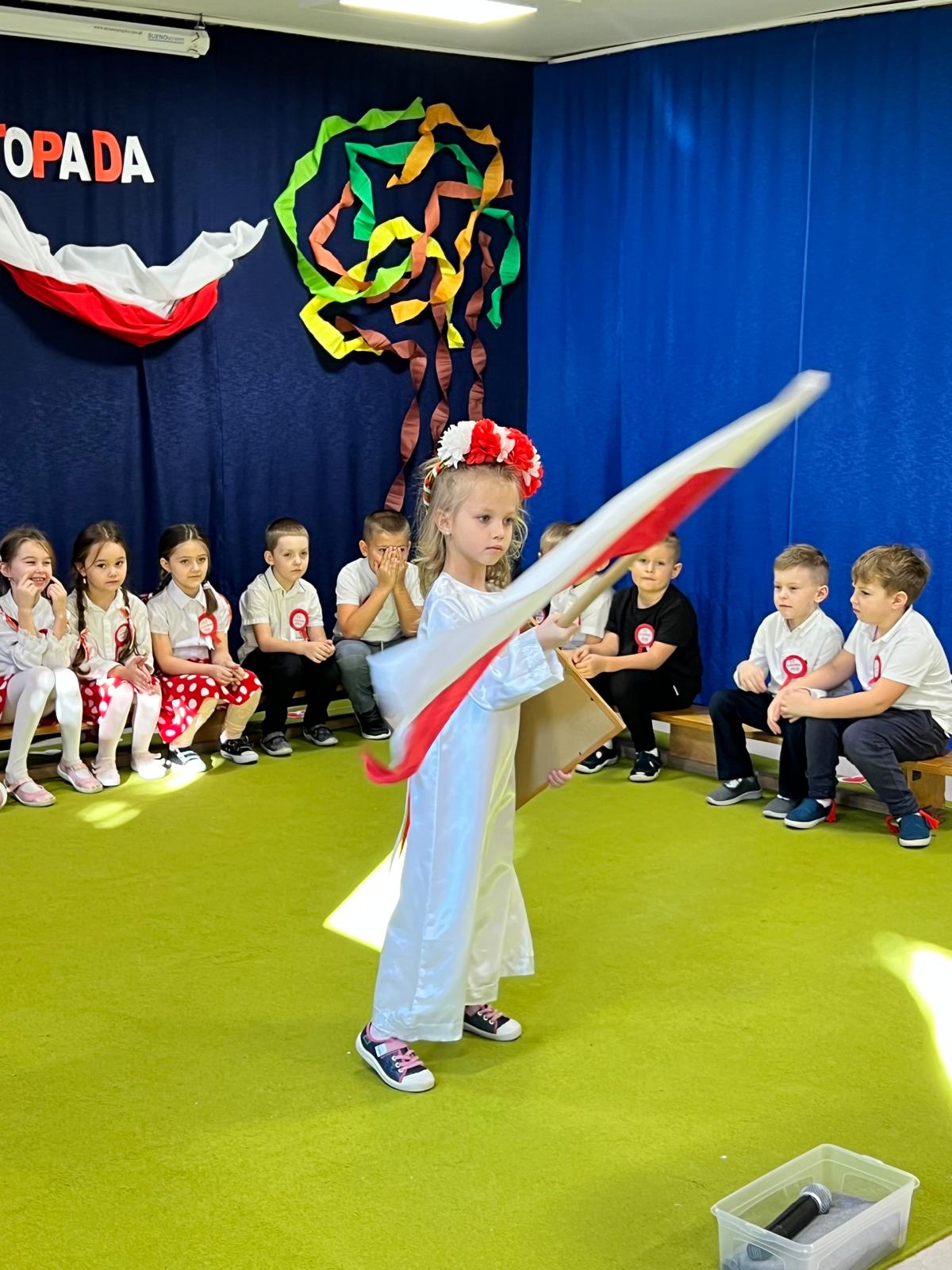 Dziewczynka ubrana w białą sukienkę, biało czerwony wianek macha Polską flagą.