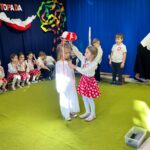 Dziewczynka ubrana w białą sukienkę otrzymuje biało czerwony wianek od dziewczynki ubranej w biała bluzkę i czerwoną spódniczkę w białe kropki.