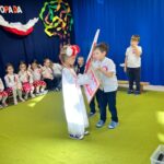 Dziewczynka ubraną w białą sukienkę otrzymuje od chłopca godło.