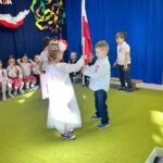 Dziewczynka ubrana w biała sukienkę otrzymuje flagę od chłopca.