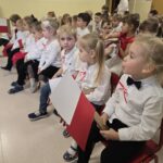 Dzieci ubrane na galowe siedzą w zali przedszkolnej podczas występu