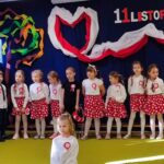 Dziewczynka w białej sukience klęczy. Za nią stoją dzieci ubrane na galowo.