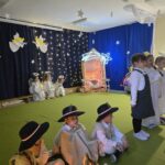 Pastuszkowie siedzą przy ognisku, w tle widać anioły i stajenkę. Trzy dziewczynki śpiewają piosenkę.