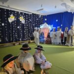 Na pierwszym planie siedzą pastuszkowie. w tle widać stajenkę przy której siedzi Maryja i Józef, a obok nich stoją anioły.
