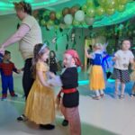 Dziewczynka przebrana za księżniczkę tańczy z chłopcem przebranym za pirata. W tle widać inne tańczące dzieci i kobietę. Wszyscy mają na sobie stroje karnawałowe