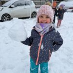 Dziewczynka w spodniach zimowych i kurtce trzyma dużą kulę śnieżną.