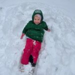 Dziewczynka w zielonej kurtce i różowych spodniach siedzi na śniegu.