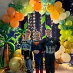 Trzech chłopców przebrani w stroje karnawałowe pozują do zdjęcia na ściance z balonów.