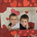Dwóch chłopców pozuje do zdjęcia trzymając czerwoną ramkę ozdobioną sercami.