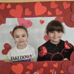Dwie dziewczynki pozują do zdjęcia trzymając czerwoną ramkę ozdobioną sercami.
