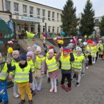 Dzieci w kamizelkach odblaskowych i kwiatkach w ręce idą na spacer.