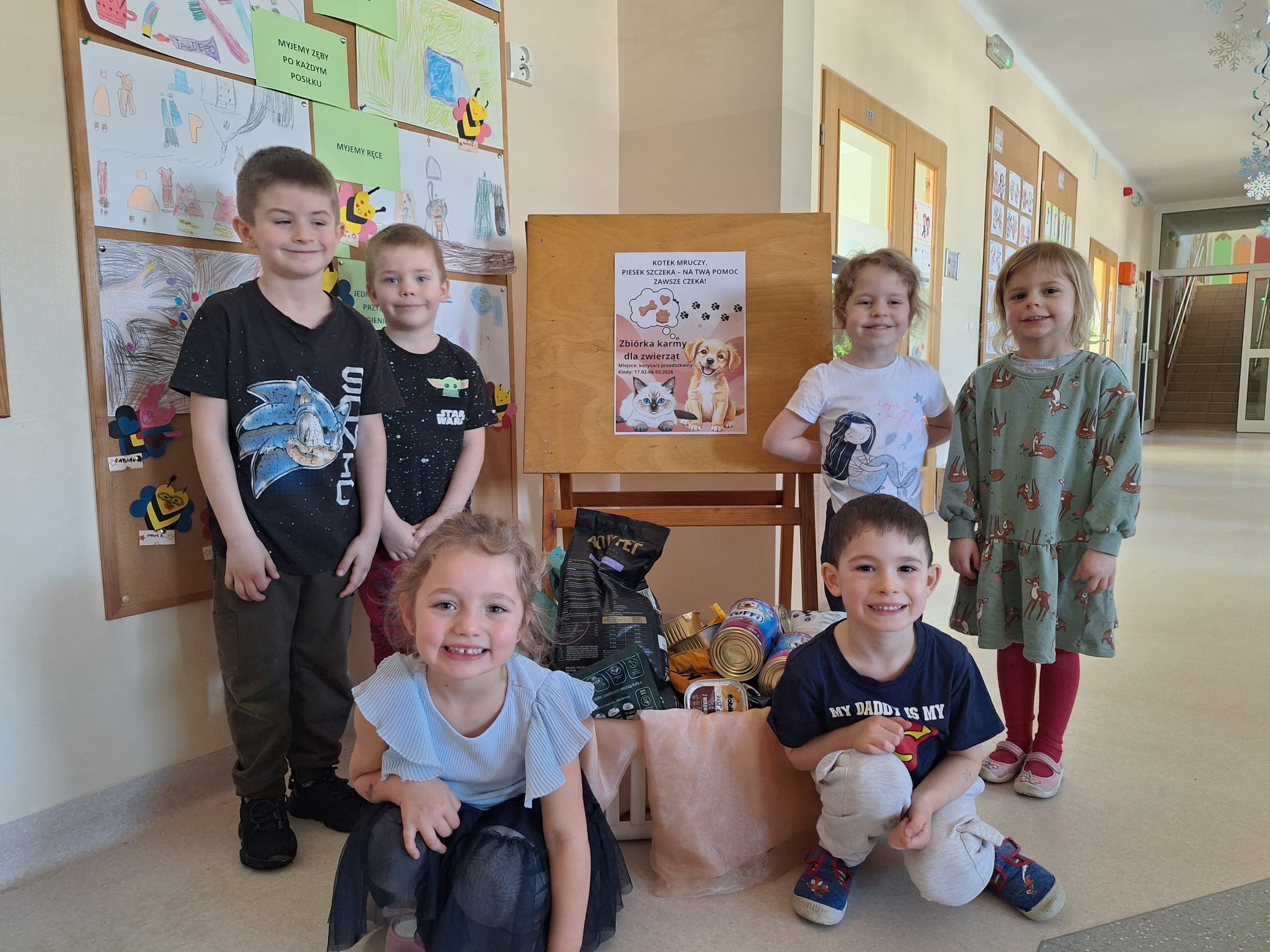 Trzy dziewczynki i trzech chłopców stoi przy koszu z karmą dla zwierząt. W tle widać plakat dotyczący zbiórki karmy dla psów i kotów.