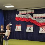 Dziewczynka i chłopiec na tle dekoracji z obchodów świąt majowych mówią wiersz.