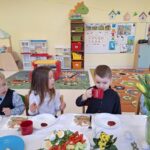 Dzieci jedzą śniadanie wielkanocne. Dziewczynka i chłopiec patrzą na chłopca pijącego herbatę.