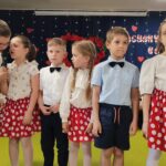 Dziewczynki w białych bluzkach i czerwonych spódniczkach w kropki oraz chłopcy ubrani elegancko recytują wiersze.