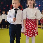 Dziewczynka w białej bluzce i czerwonej spódniczce w kropki oraz chłopiec ubrany elegancko recytują wiersze.