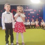 Dziewczynka w białej bluzce i czerwonej spódniczce w kropki oraz chłopiec ubrany elegancko recytują wiersze.