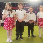 Dziewczynki w białych bluzkach i czerwonych spódniczkach w kropki oraz chłopcy ubrani elegancko recytują wiersze.