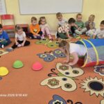 Dzieci w sali przedszkolnej siedzą na dywanie a dziewczynka przechodzi przez tunel.