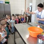 Na zdjęciu widzimy grupkę dzieci w wieku przedszkolnym zebrała się wokół kuchni, uważnie słuchając instrukcji szefa kuchni.