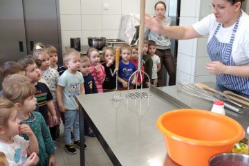 Na zdjęciu widzimy grupkę dzieci w wieku przedszkolnym zebrała się wokół kuchni, uważnie słuchając instrukcji szefa kuchni.