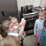 Na zdjęciu widzimy grupę dzieci w wieku przedszkolnym gromadzi się wokół kuchni podczas lekcji gotowania.